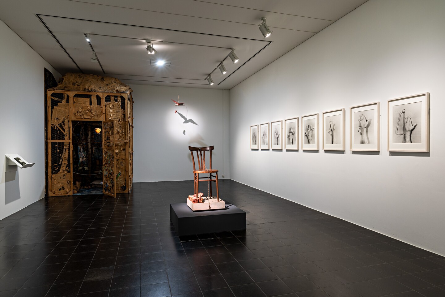 Galerieraum mit gerahmten Werken an weißer Wand, Skulptur in der Mitte und Installation hinten rechts