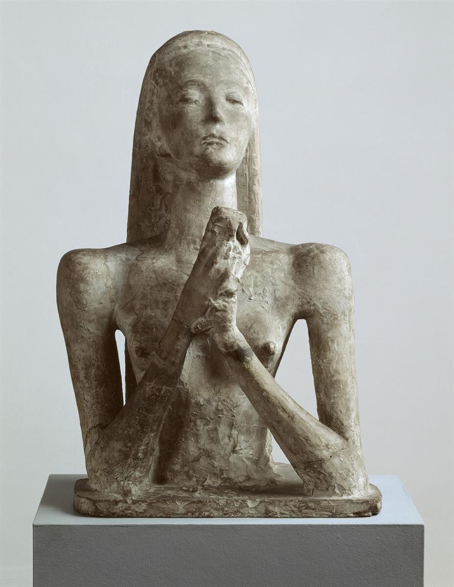 Steinskulptur einer sitzenden Figur mit betenden Händen auf grauem Sockel