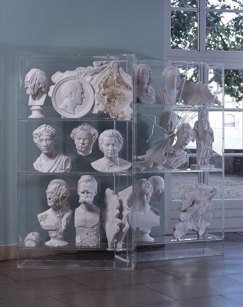 Kunstinstallation mit gestapelten menschlichen Büsten und Köpfen aus hellem Material in zerschlagenen Vitrinen