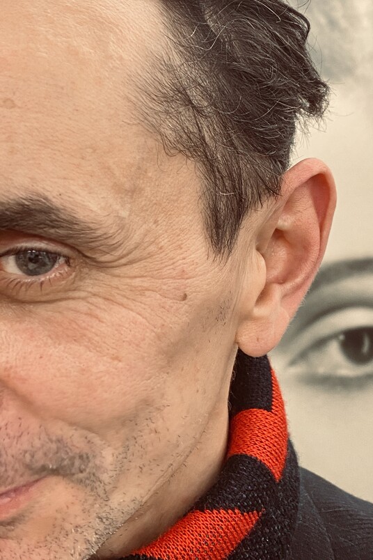 Nahaufnahme einer Gesichtshälfte eines Mannes mit schwarzem Haar und rot-schwarzem Schal. Im Hintergrund ein unscharfes schwarz-weiß Portrait einer Frau, deren Auge gut erkennbar ist.