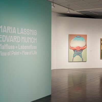 Galerieraum mit türkiser Wand, Text und zwei gerahmten Kunstwerken an weißer Wand